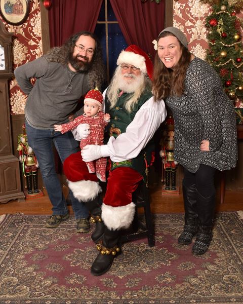 Parents with their baby in a red knit hat, sitting on Santa’s knee